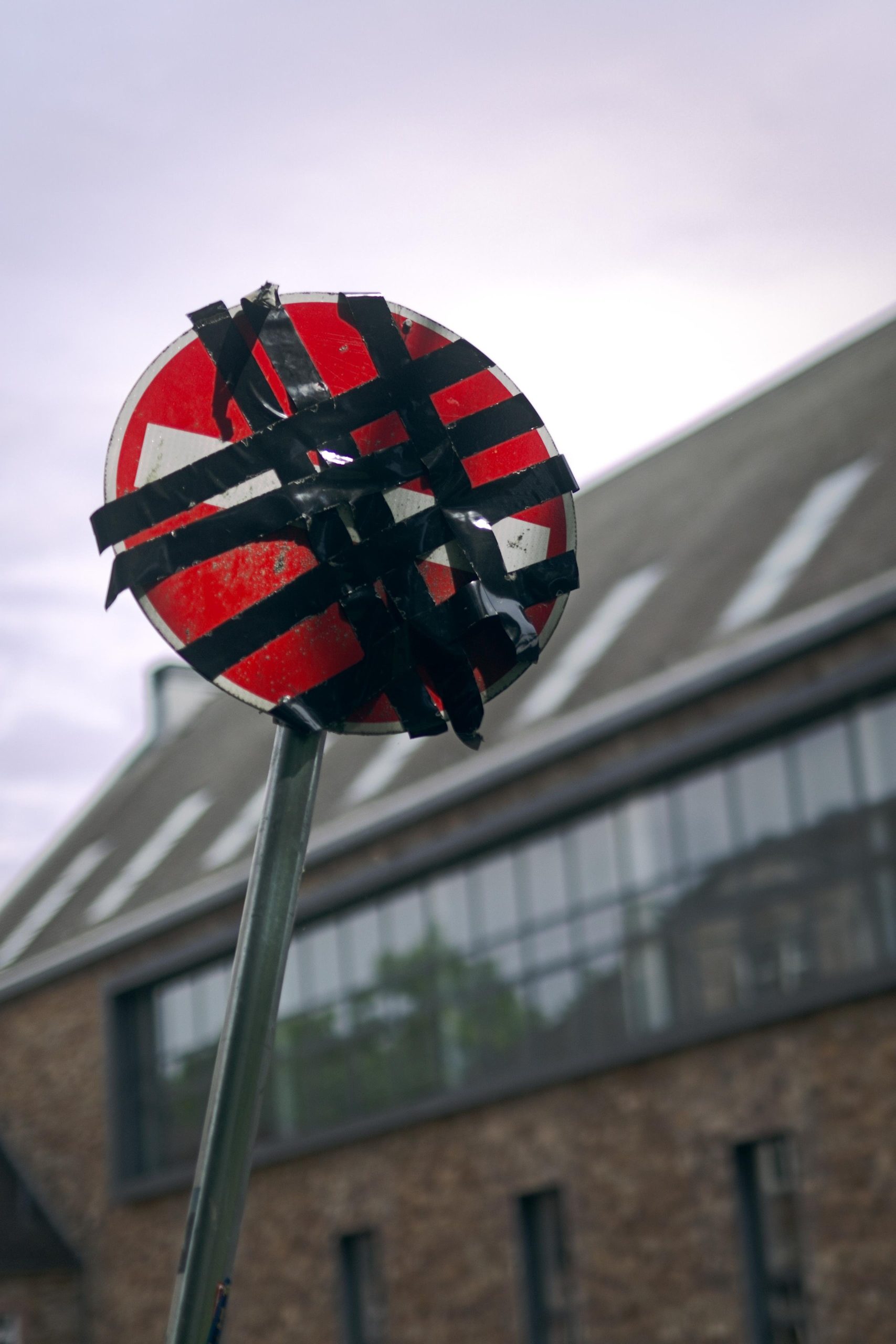 Red circular street sign with a white horizontal bar, covered with overlapping strips of black tape, mounted on a pole against an overcast sky with a blurred building in the background.