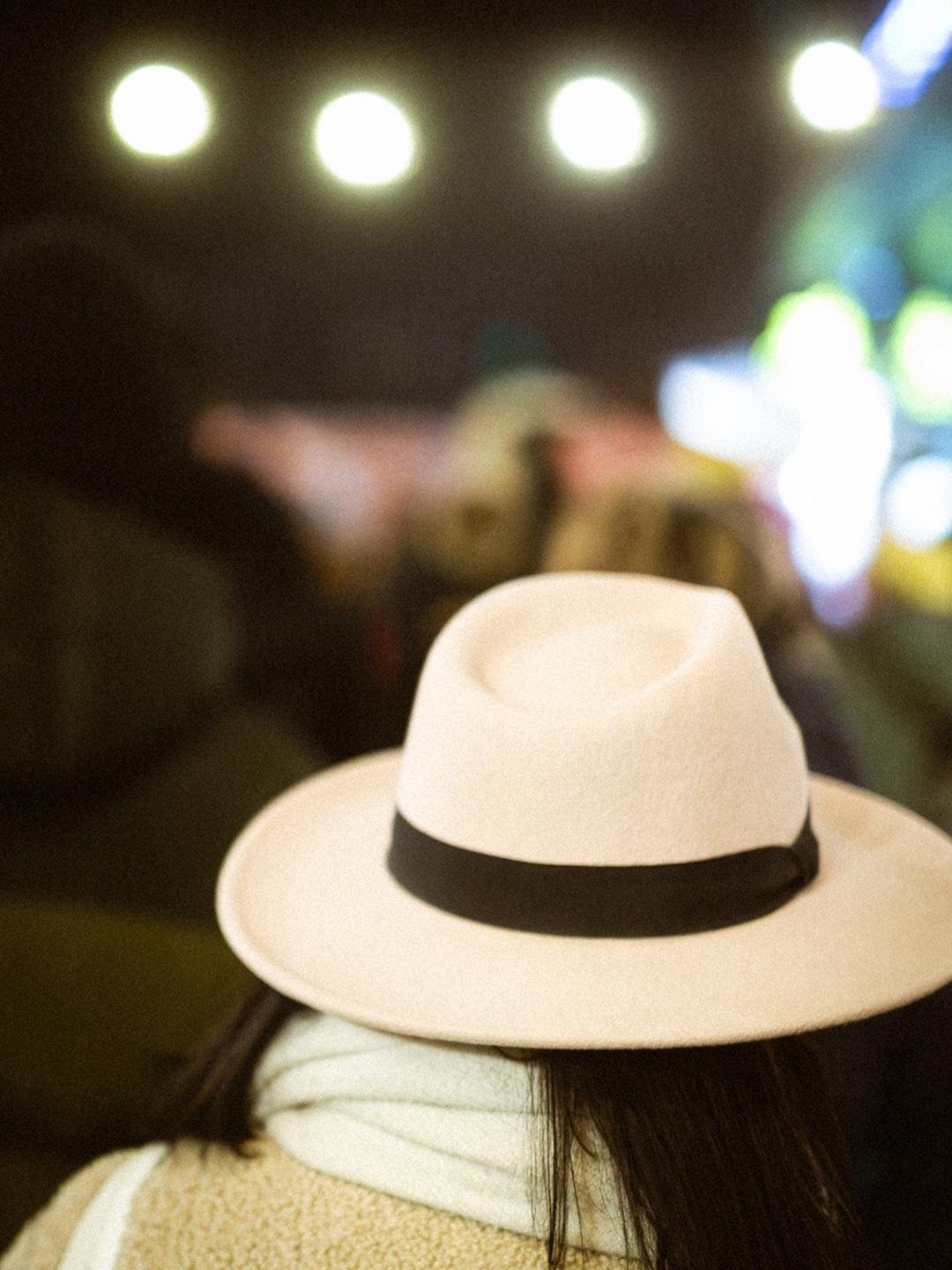 Rear view of a person wearing a beige wide-brim hat with a black band and a light scarf, with blurred nighttime lights in the background and three bright circular lights along the top.
