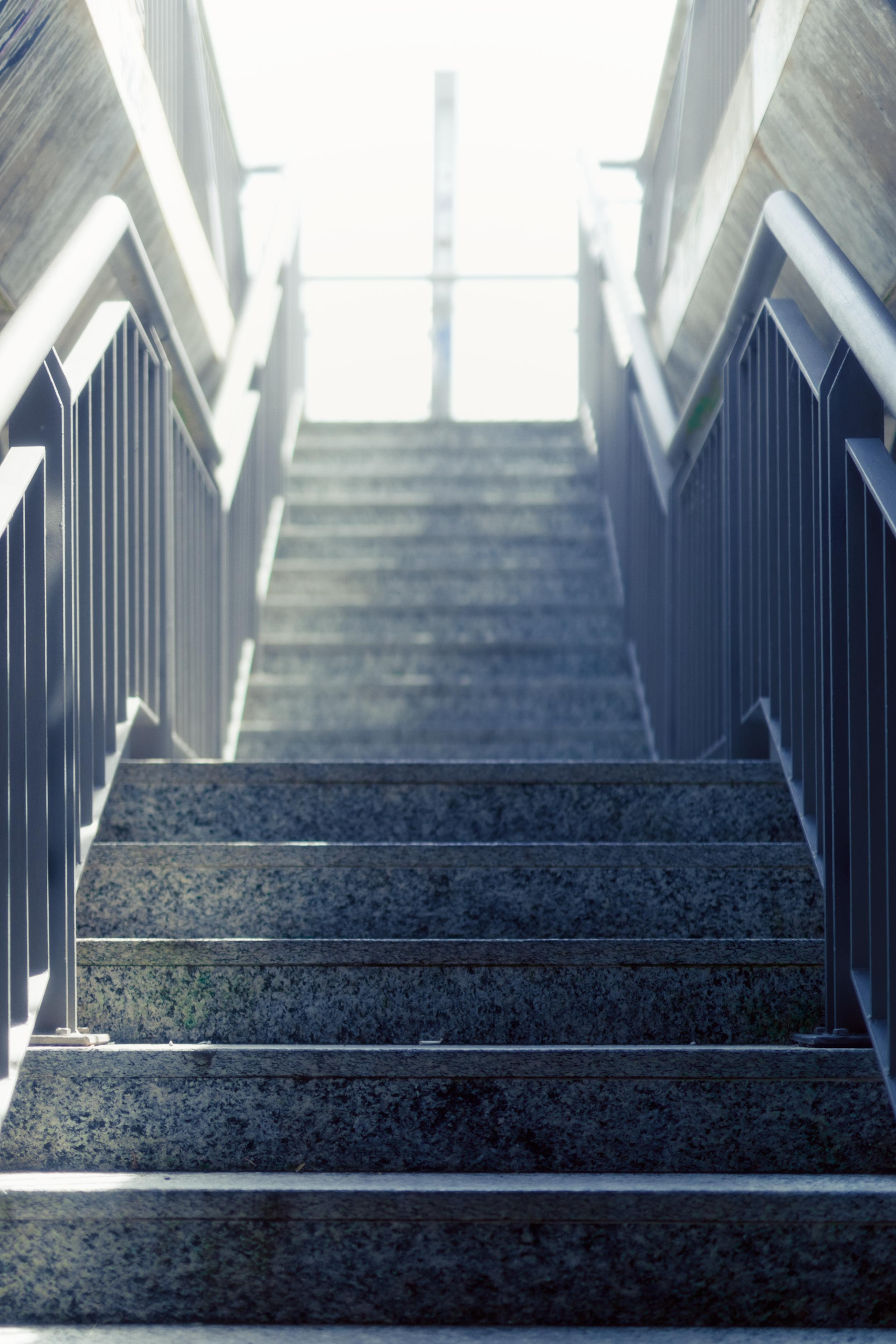 Upward view of a concrete stairway with metal railings on both sides, leading to an overexposed bright opening at the top.