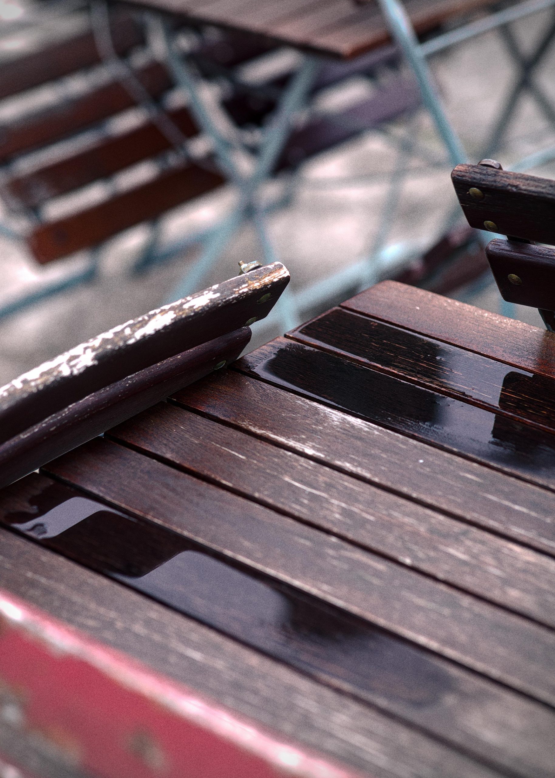 Close-up of a wet wooden outdoor table with puddles and reflections on the slats, with blurred metal-framed chairs and benches in the background.