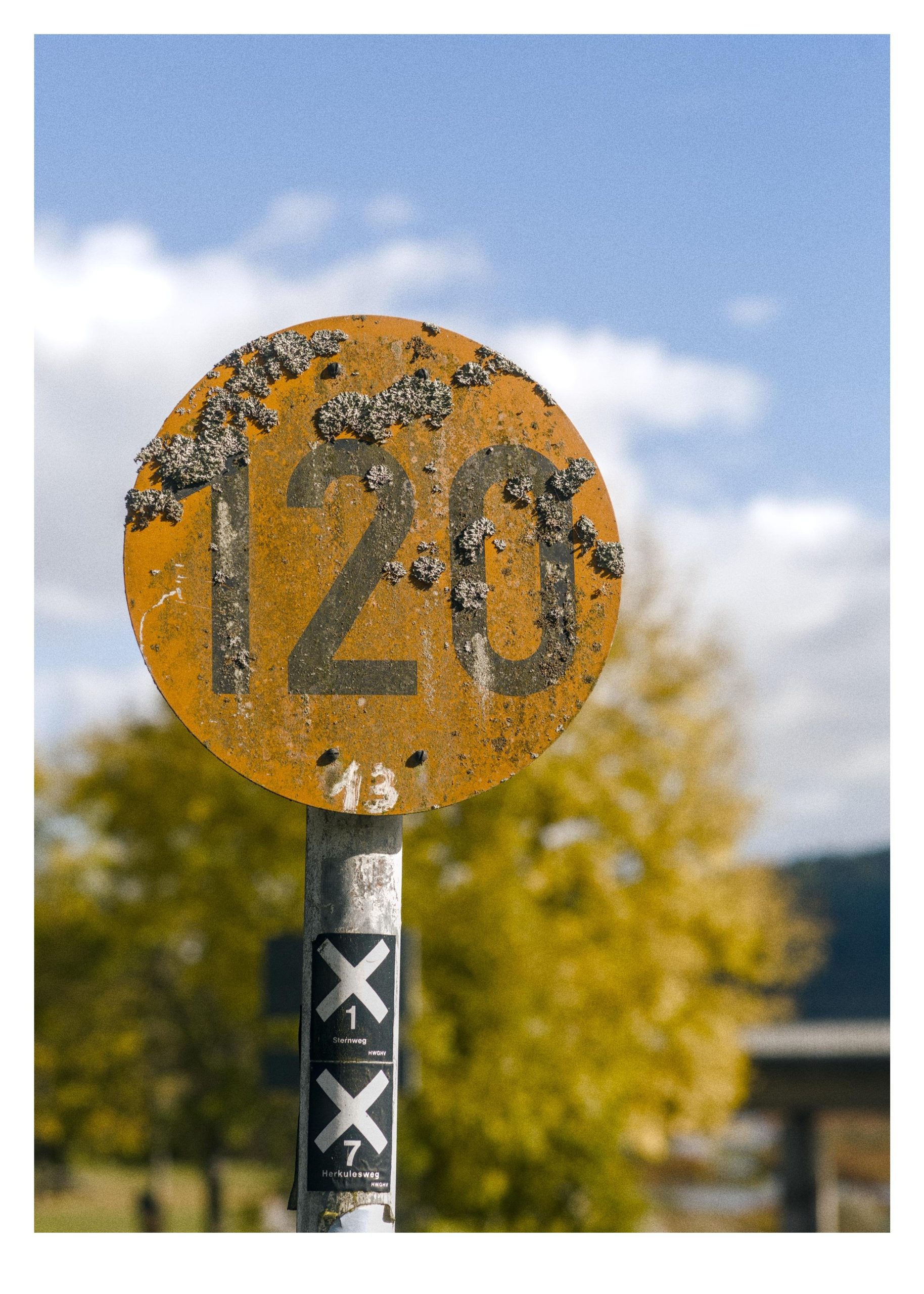 Weathered circular yellow sign with the number “120” in black, speckled with lichen, mounted on a metal pole with two black-and-white “X” stickers; blurred trees and a blue sky with clouds in the background.