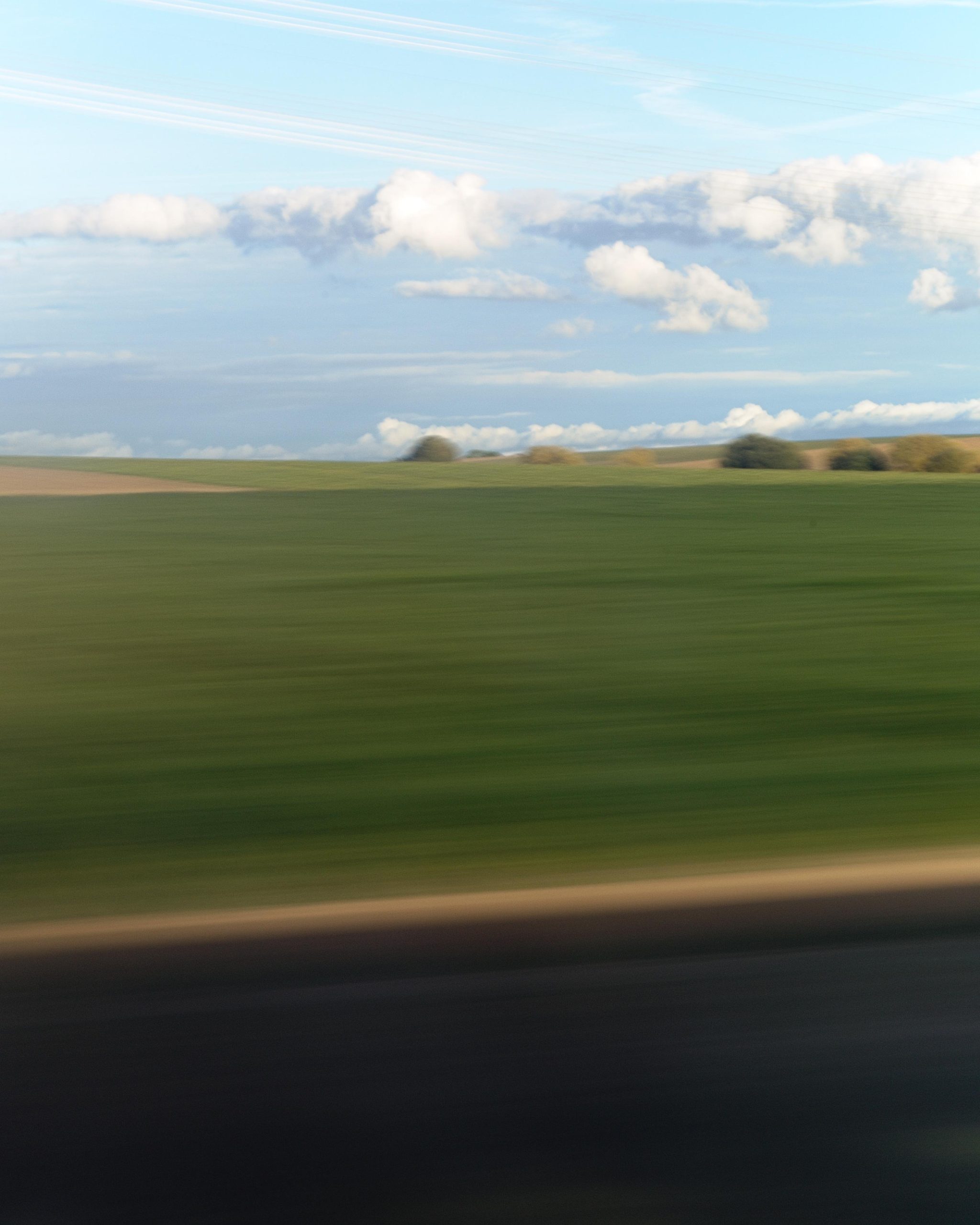 Blurred view of green farmland and a distant tree line under a blue sky with scattered white clouds, with motion blur streaks across the lower edge and foreground.