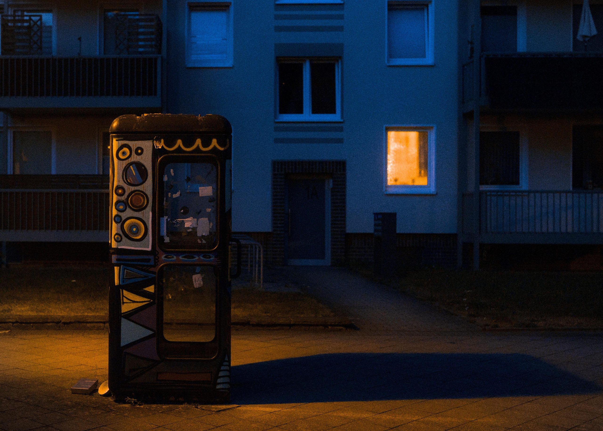 Graffiti-painted phone booth on a dimly lit sidewalk at night, with a dark apartment building behind it and one warmly lit window on the right casting light onto the pavement.