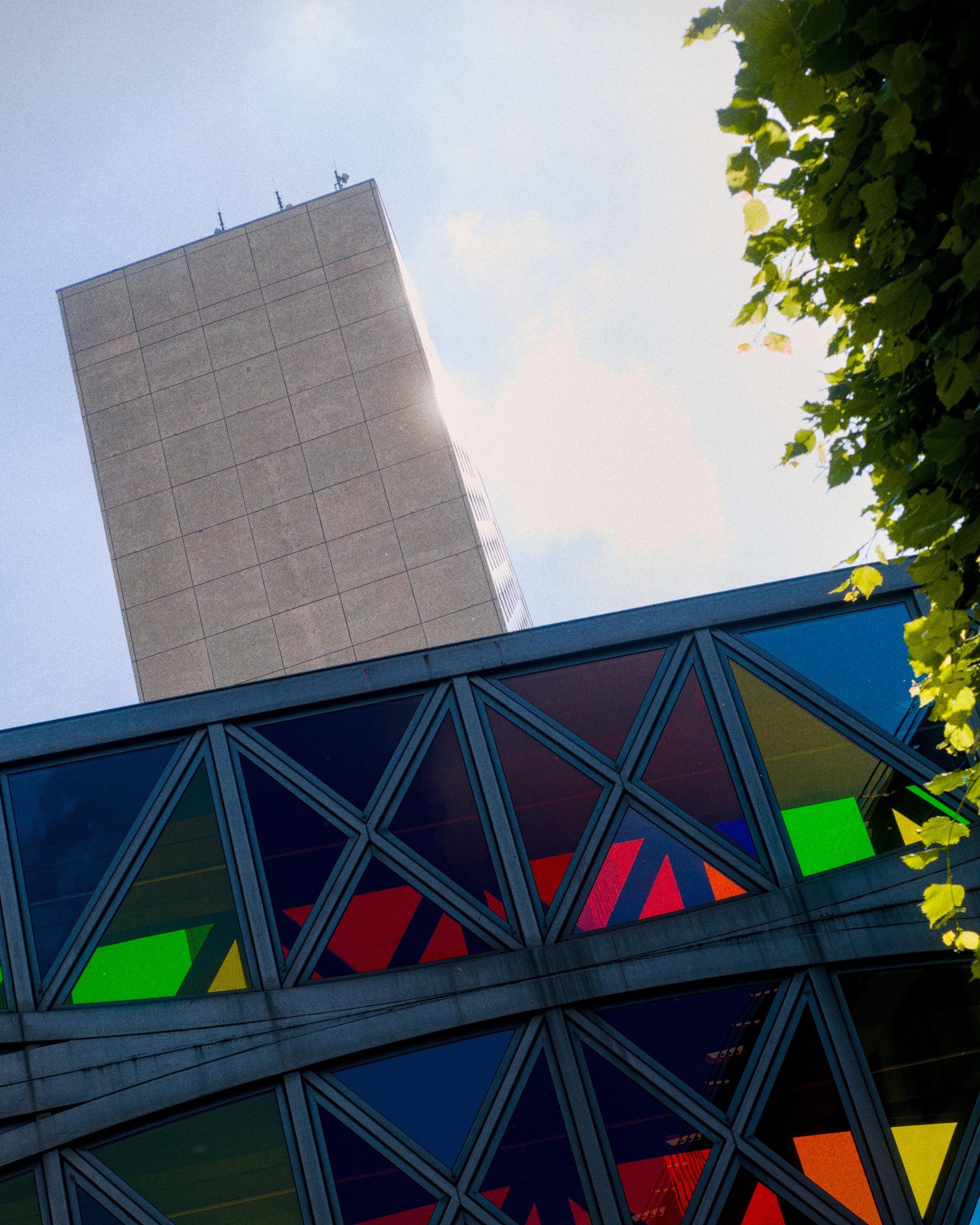 Low-angle view of a tall concrete tower behind a modern building facade with dark crisscross metal framing and colorful glass panels in green, red, blue, and yellow; leafy branches frame the upper right against a pale blue sky with clouds.
