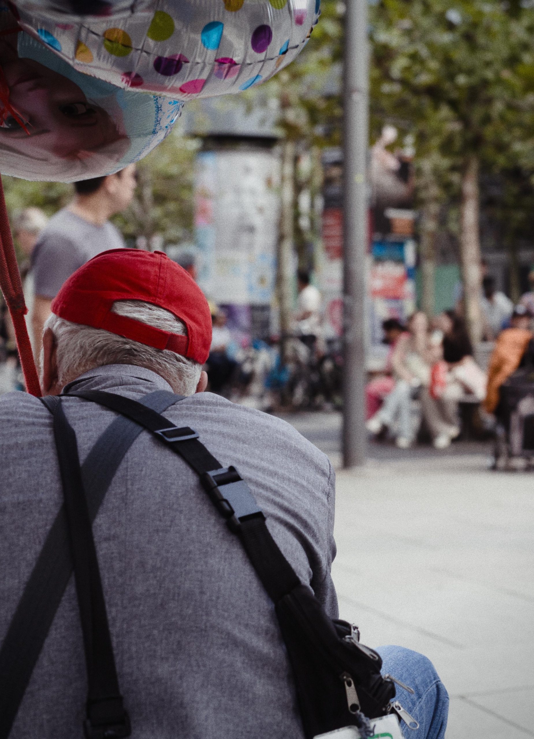 Rear view of an older person wearing a red cap and gray shirt with a black backpack strap, seated on a city sidewalk; a polka-dot ballon and a partial face appear at the top left, with pedestrians and benches blurred in the background.