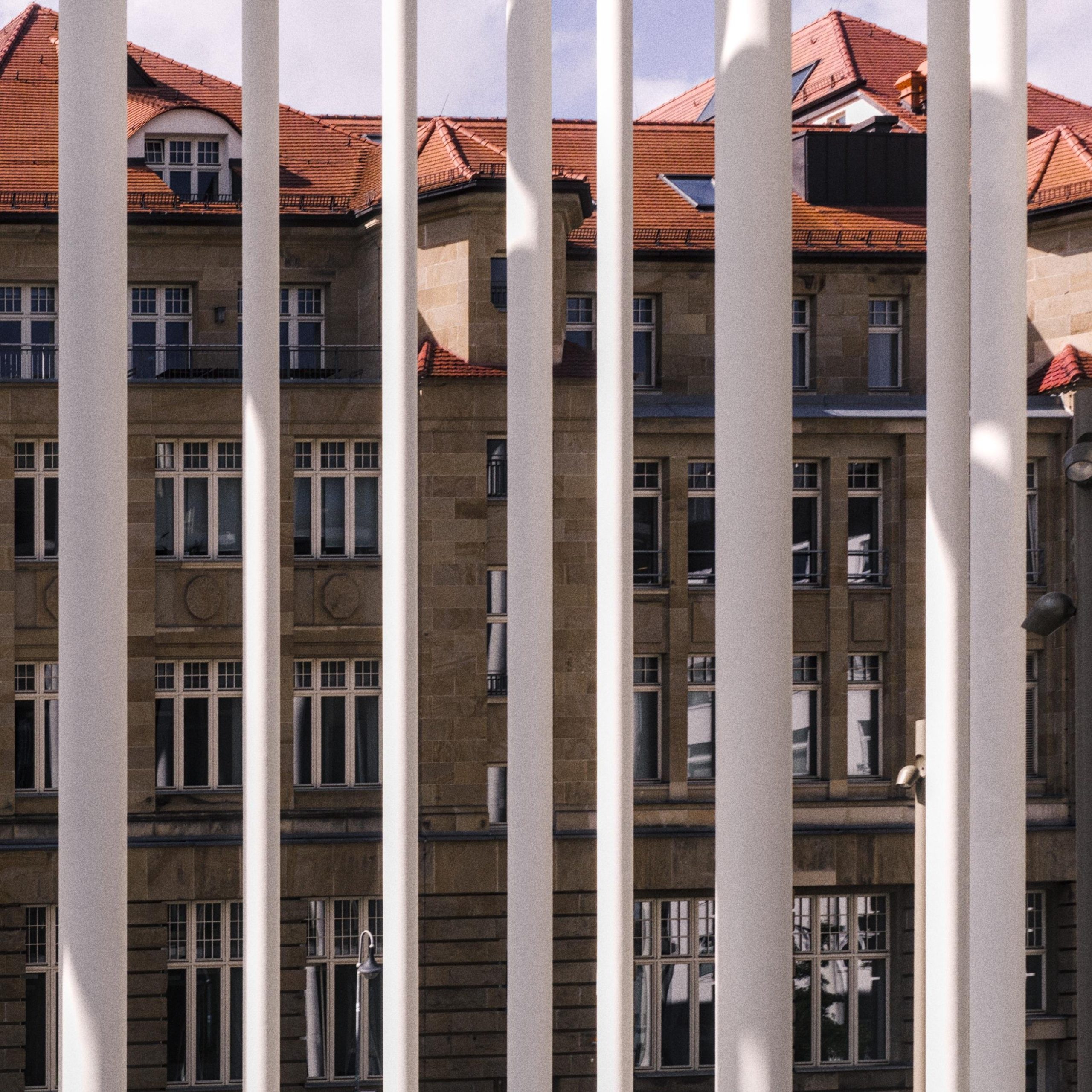 Tall white vertical bars in the foreground partially obscure a multi-story stone building with many rectangular windows and red-tiled rooftops under a pale sky.