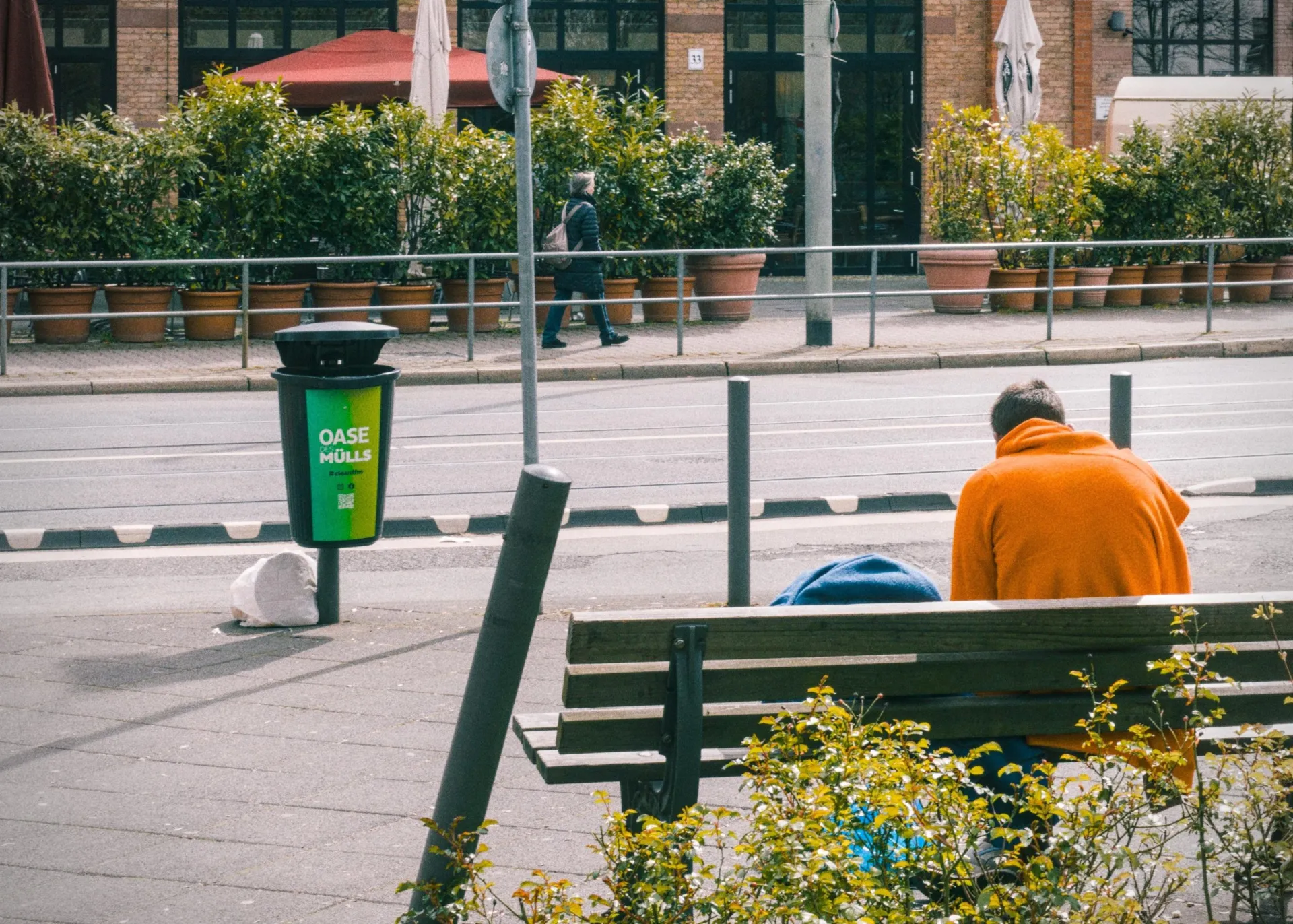 Person in an orange jacket sitting on a wooden bench beside a bundled blue item, with a green “OASE MÜLLS” trash bin nearby; across the street are potted shrubs, a metal railing, and a brick building entrance in the background.