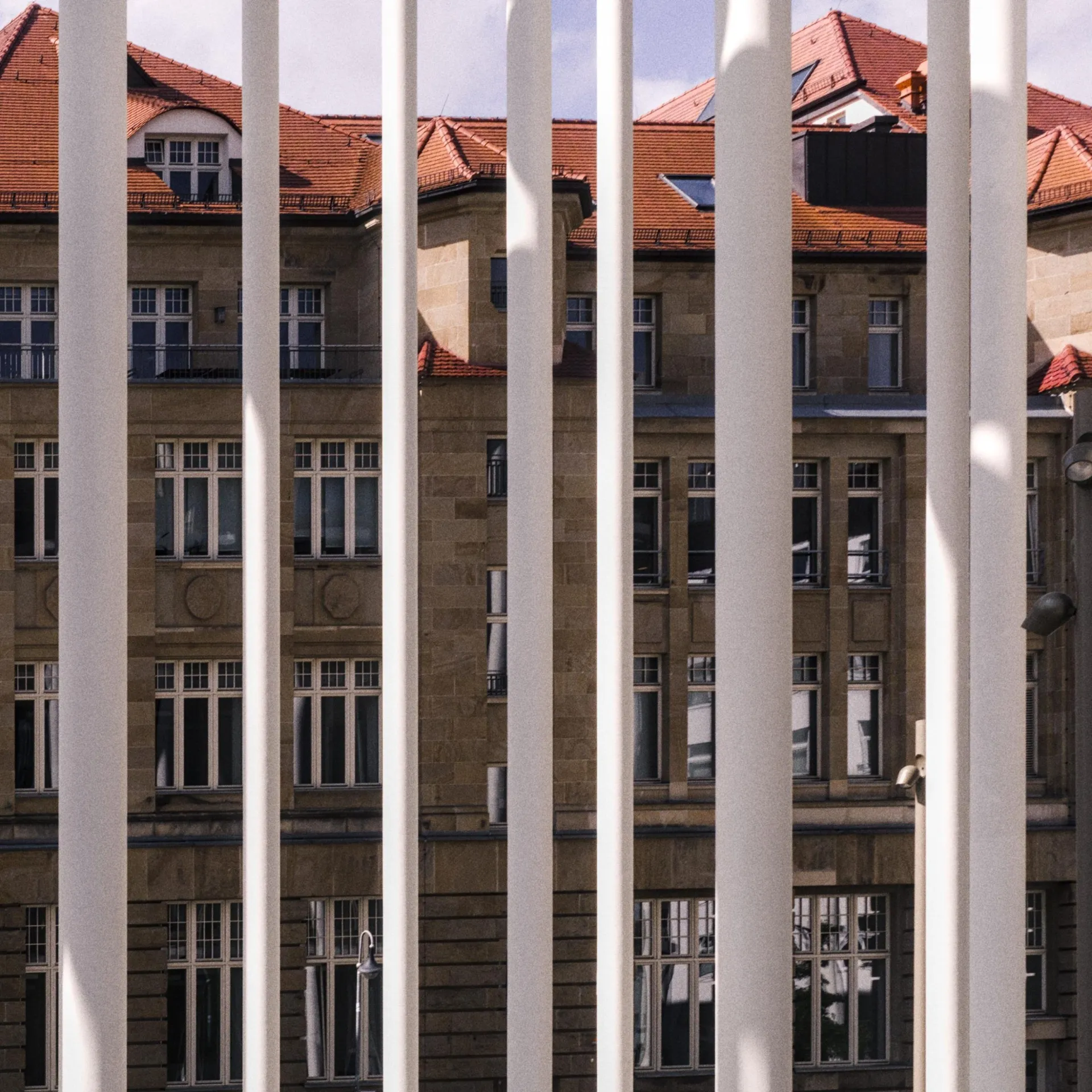 Tall white vertical bars in the foreground partially obscure a multi-story stone building with many rectangular windows and red-tiled rooftops under a pale sky.