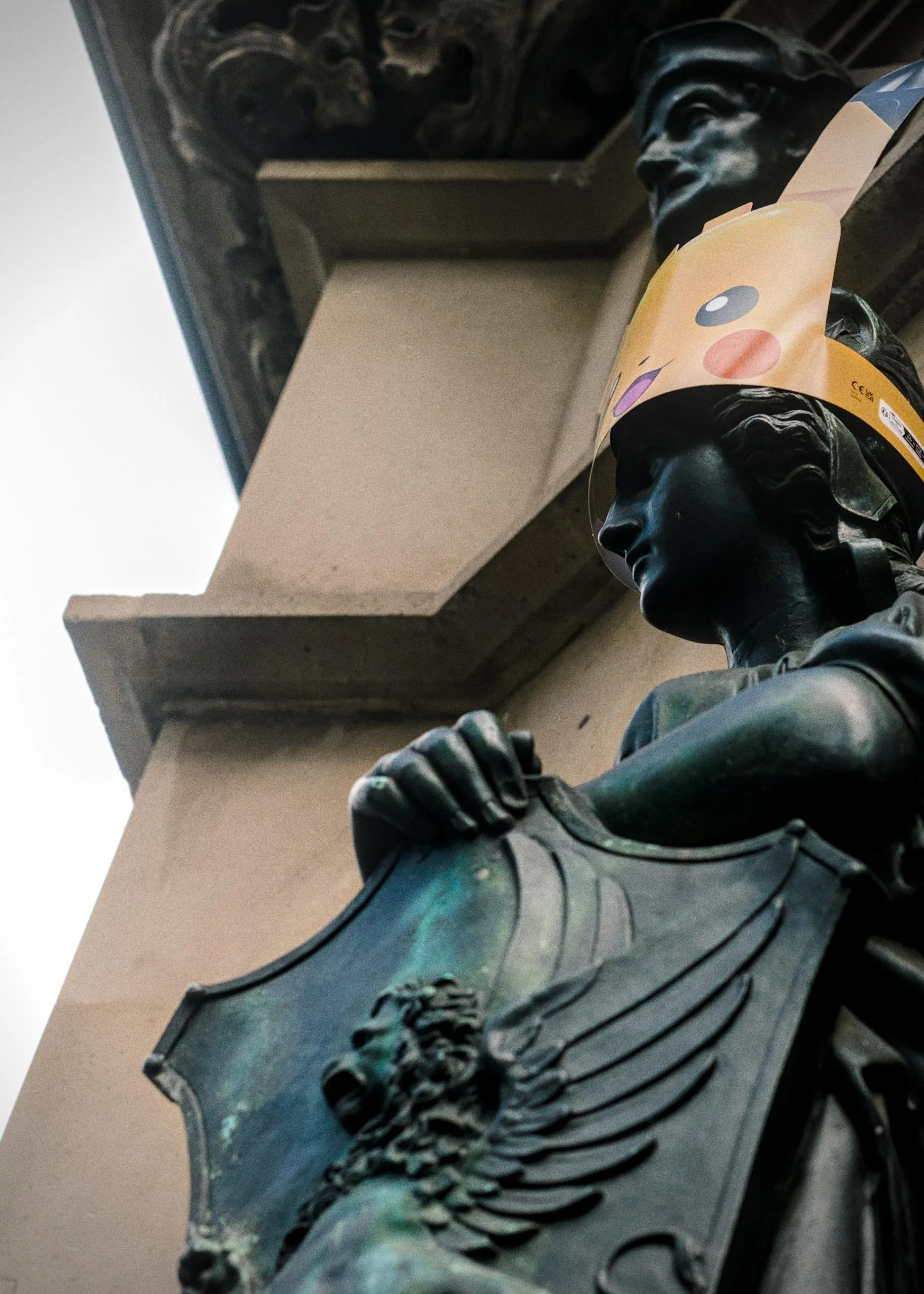 Bronze statue on a building facade holding a large shield, with a paper Pikachu face mask taped over the statue’s head; shot from below with stone columns and the underside of an overhang in the background.