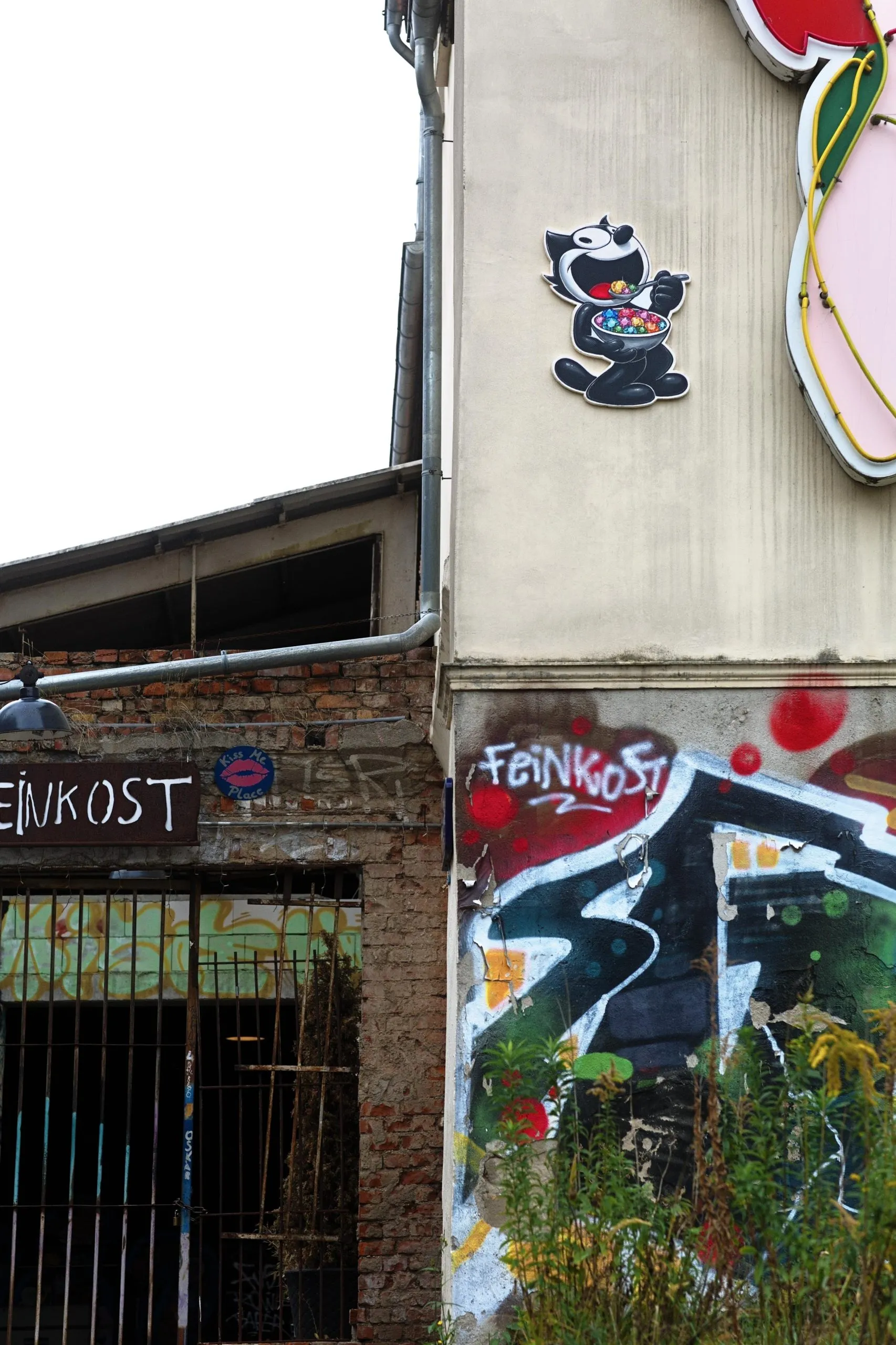 Exterior wall of an urban building with graffiti and the word “FEINKOST” painted in white, a small Felix the Cat sign holding a bowl mounted high on the wall, a barred doorway and brickwork on the left, and overgrown weeds at the base.