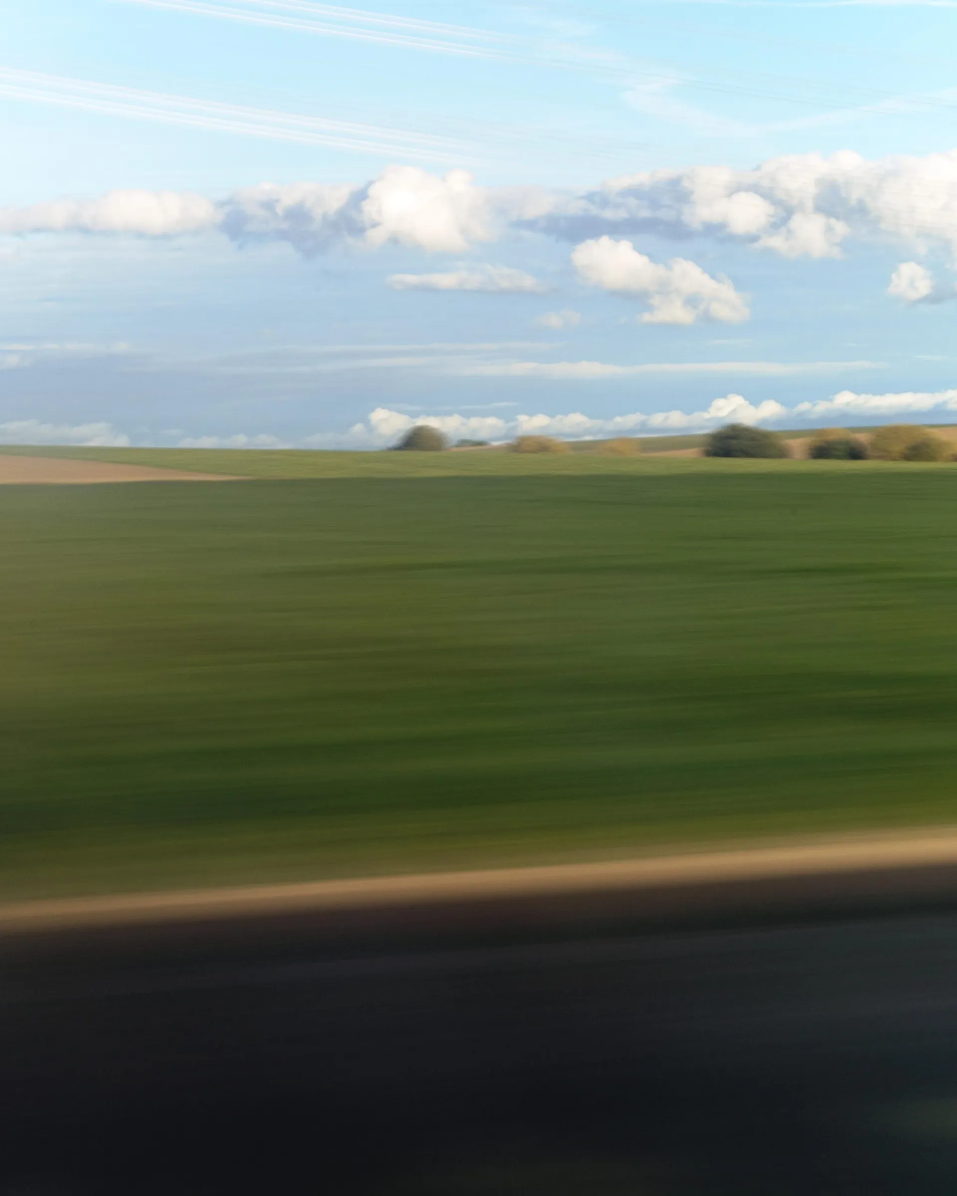 Blurred view of green farmland and a distant tree line under a blue sky with scattered white clouds, with motion blur streaks across the lower edge and foreground.